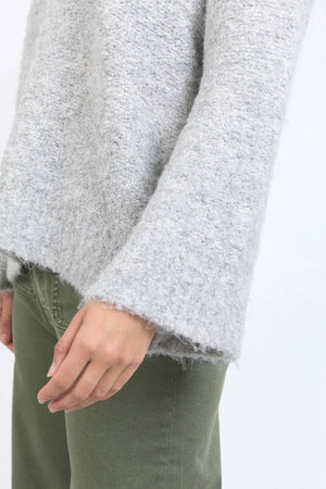 Wearing the Foxwood Mindie Knit Crew, a fuzzy light gray sweater with wide sleeves and relaxed fit, paired with olive green pants, a person is shown from the shoulders down with their hand relaxed at their side.