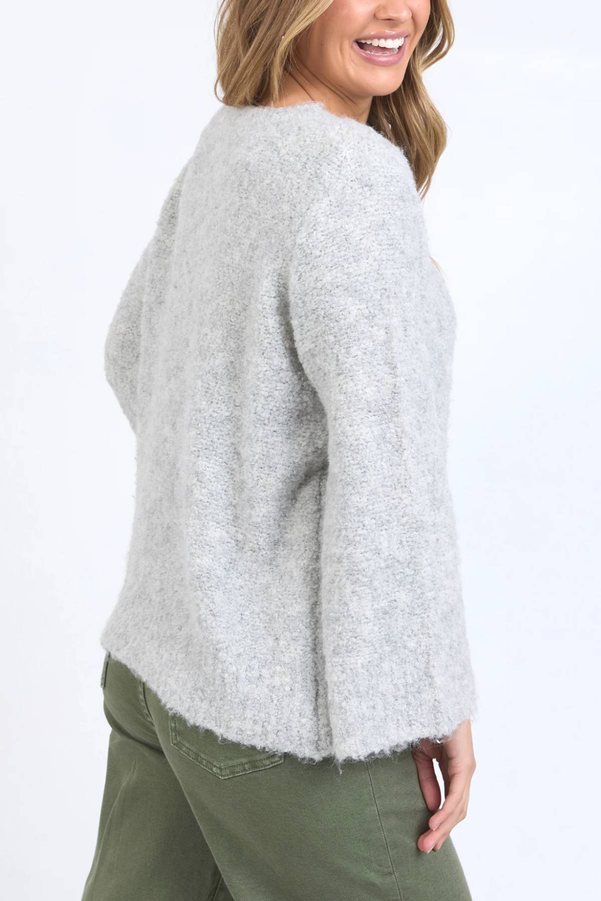 A woman smiles in the Foxwood Mindie Knit Crew, a relaxed-fit light gray sweater with loose sleeves, paired with olive green pants and set against a plain white background.