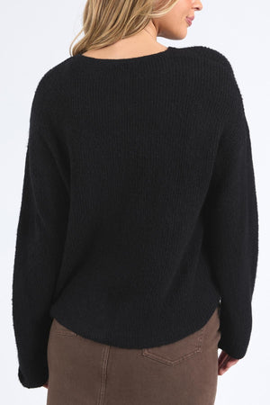 A woman faces away from the camera, showcasing the Foxwood Murphy Knit Cardi in black—a relaxed fit sweater with ribbed trims—paired with brown pants, against a plain light background.