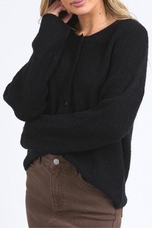 A woman wearing the Foxwood Murphy Knit Cardi in black with ribbed trims and brown pants crosses her arms, one hand touching her neck, against a plain white background.