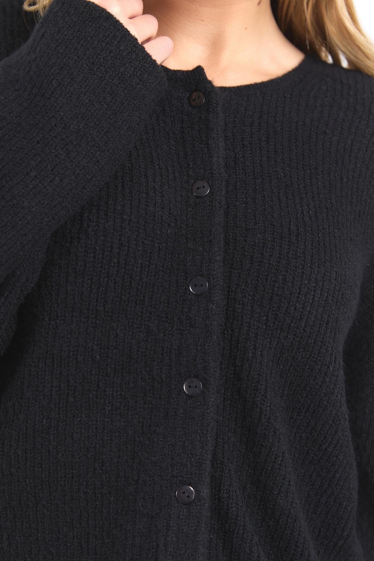 A person wears the Foxwood Murphy Knit Cardi in black, featuring long sleeves, black front buttons, and ribbed trims. Only their upper torso and part of their face are visible.