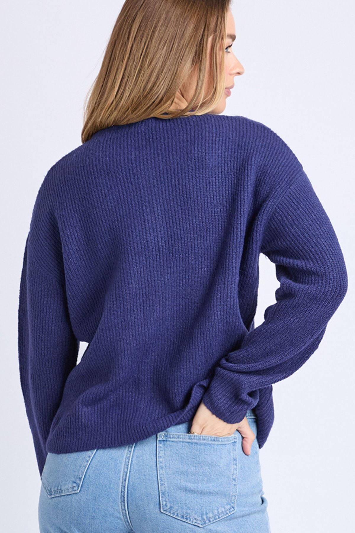A woman with light brown hair, seen from behind, wears a loose-fitting Foxwood Murphy Knit Cardi in dark blue and light blue jeans. She stands against a plain light background with one hand in her pocket.