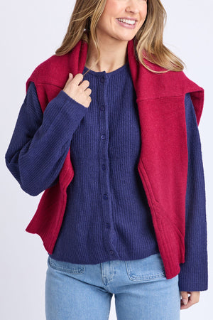 A woman smiles wearing the Foxwood Murphy Knit Cardi in blue, paired with light blue jeans and a red sweater draped over her shoulders, set against a plain light background.