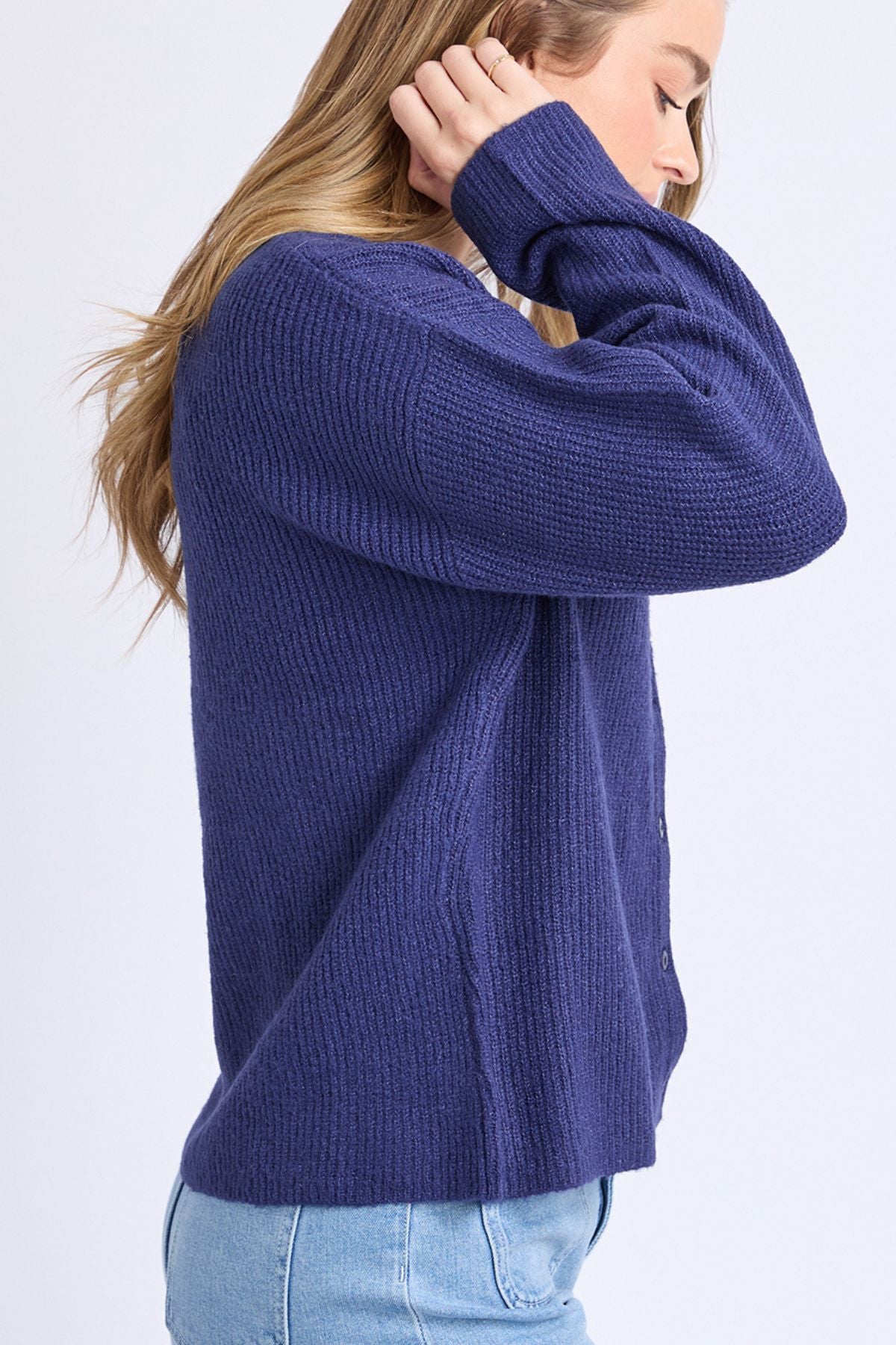 A person with long, light brown hair wears a chunky, ribbed Foxwood Murphy Knit Cardi in dark blue and light blue jeans, shown in side profile with one hand raised to their head against a plain, light background.
