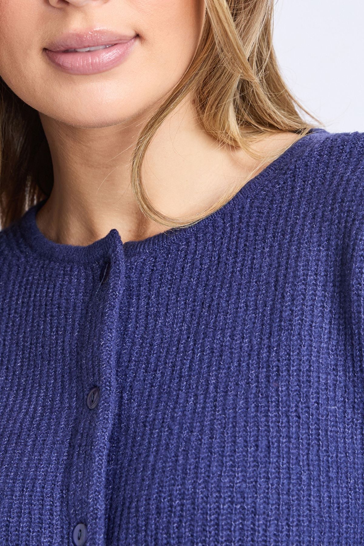 A close-up shows a woman wearing the Foxwood Murphy Knit Cardi, with only her lower face, neck, and upper chest visible. Her lips are slightly parted, and soft light brown hair falls over the textured dark blue cardigan.