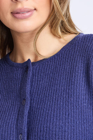 A close-up shows a woman wearing the Foxwood Murphy Knit Cardi, with only her lower face, neck, and upper chest visible. Her lips are slightly parted, and soft light brown hair falls over the textured dark blue cardigan.