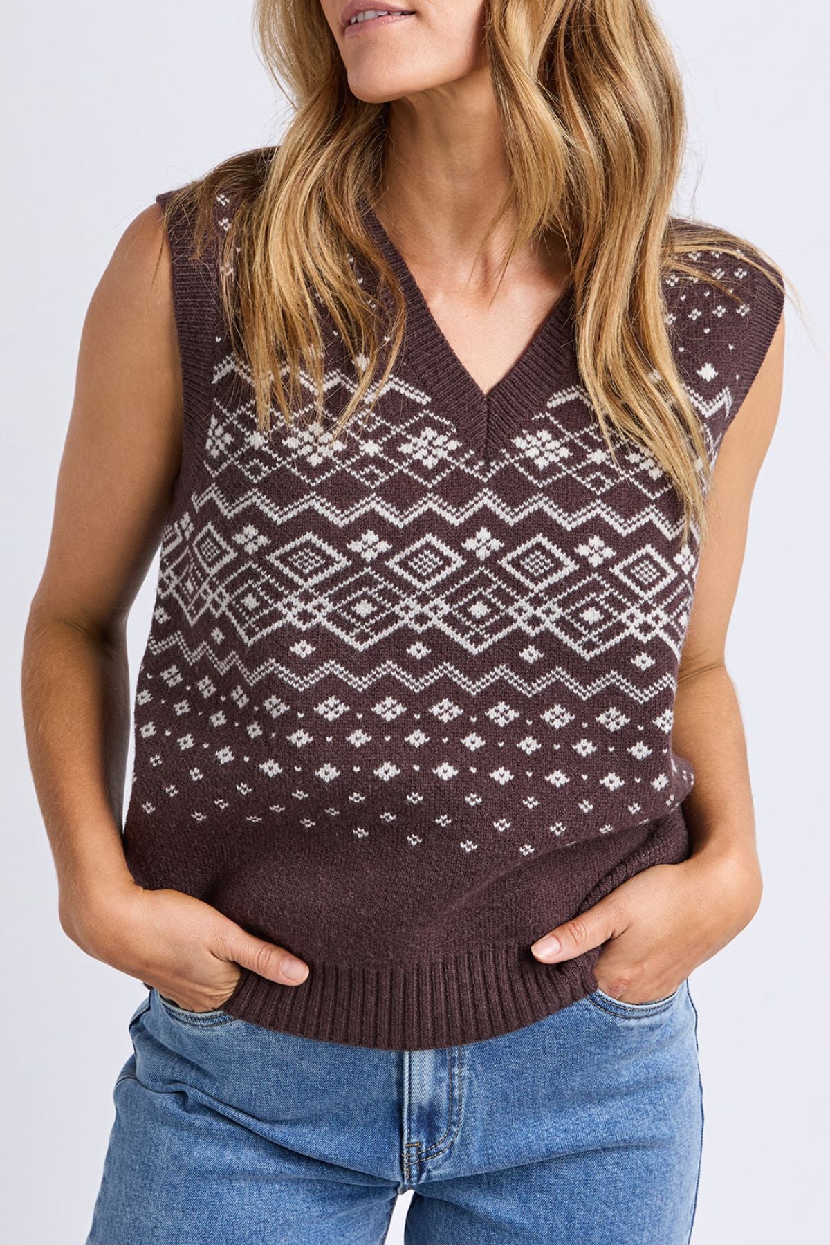A woman with wavy light brown hair wears the Foxwood Nordic Vest, a brown knitted sleeveless sweater with a white geometric pattern, paired with blue jeans and her hands in her pockets.