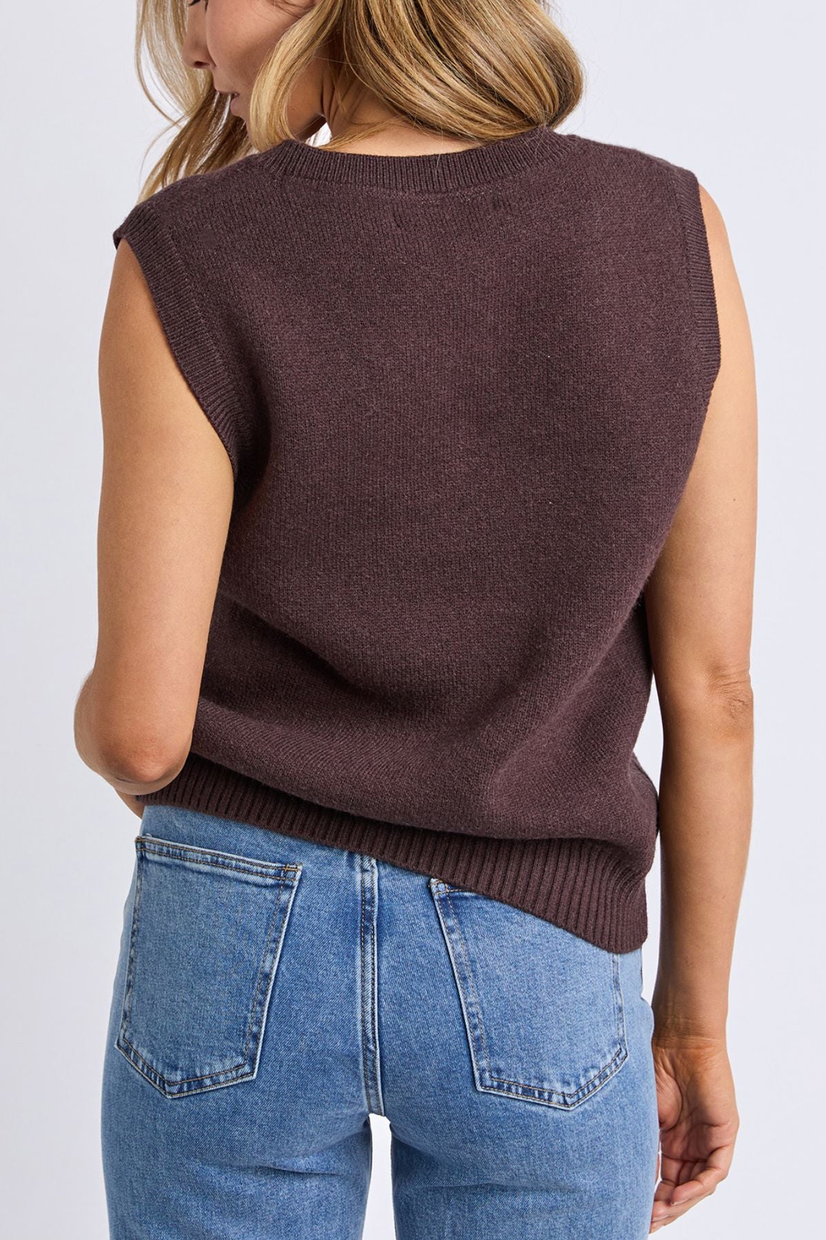 A woman with wavy blonde hair stands with her back to the camera against a plain light background, wearing the Foxwood Nordic Vest and high-waisted blue jeans.