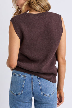 A woman with wavy blonde hair stands with her back to the camera against a plain light background, wearing the Foxwood Nordic Vest and high-waisted blue jeans.
