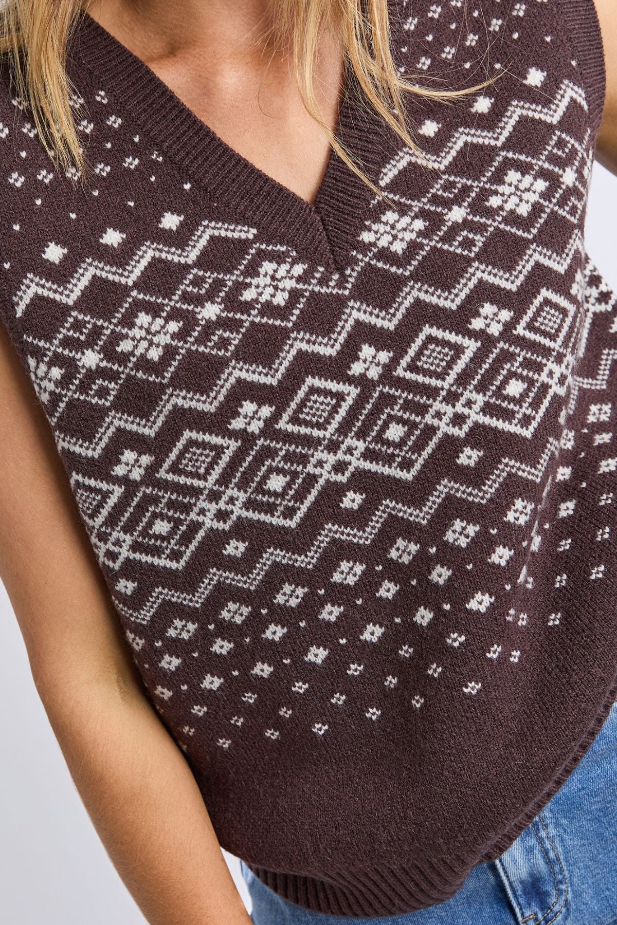 Someone wearing the Foxwood Nordic Vest, a brown knit with a white geometric and snowflake pattern, paired with blue jeans. The image is cropped to show the torso and part of one arm.