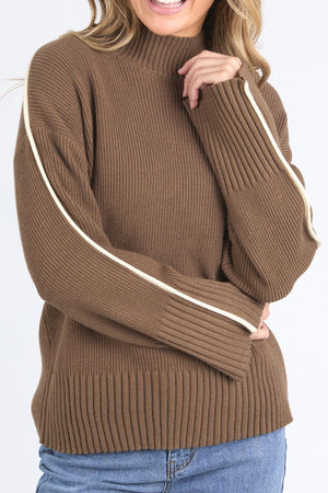A person wears the Foxwood Oliver Knit—a ribbed brown turtleneck sweater with cream stripes on the sleeves—paired with light blue jeans, smiling and casually touching their hair.