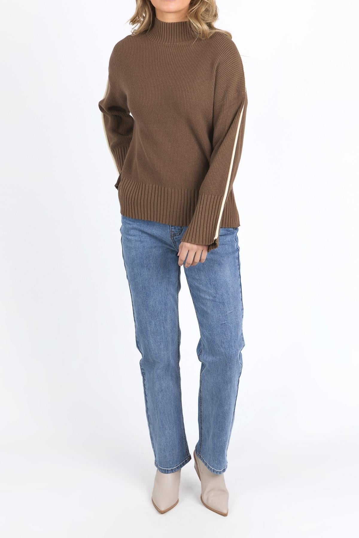 A person wears the Foxwood Oliver Knit—a brown, relaxed-fit ribbed sweater with white sleeve stripes—paired with blue jeans and beige pointed-toe ankle boots, standing against a plain white background.