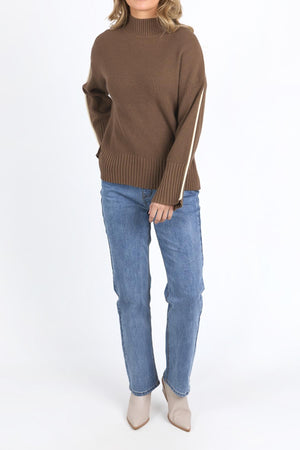 A person wears the Foxwood Oliver Knit—a brown, relaxed-fit ribbed sweater with white sleeve stripes—paired with blue jeans and beige pointed-toe ankle boots, standing against a plain white background.