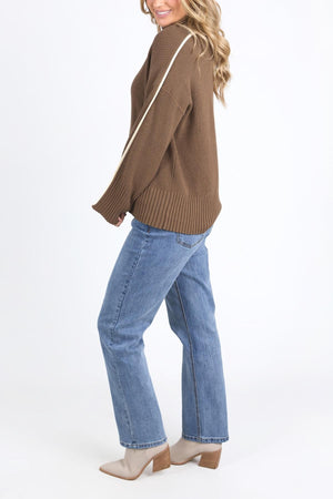 A woman stands sideways, smiling, wearing the Foxwood Oliver Knit sweater with a white sleeve stripe, blue jeans, and taupe heeled ankle boots against a plain white background.