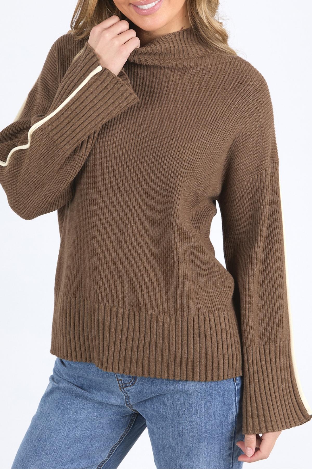 A woman wearing the Foxwood Oliver Knit, a brown ribbed sweater with wide sleeves, white arm stripe, and mock neck, pairs it with blue jeans and smiles while holding the collar near her chin for a relaxed look.