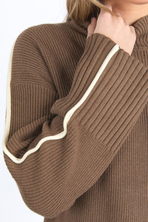 A person is shown from the shoulders down wearing the Foxwood Oliver Knit, a brown ribbed sweater with a cream stripe on the sleeve and a mock neck, with one hand touching the collar.