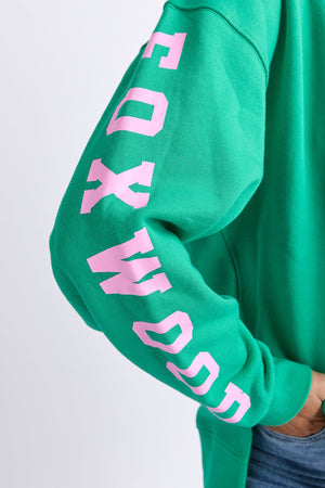 A person wears a Foxwood Origin Crew in bright green, featuring bold pink "FOXWOOD" lettering down the sleeve. Their hand is on their hip and they pair it with blue jeans for a modern look.