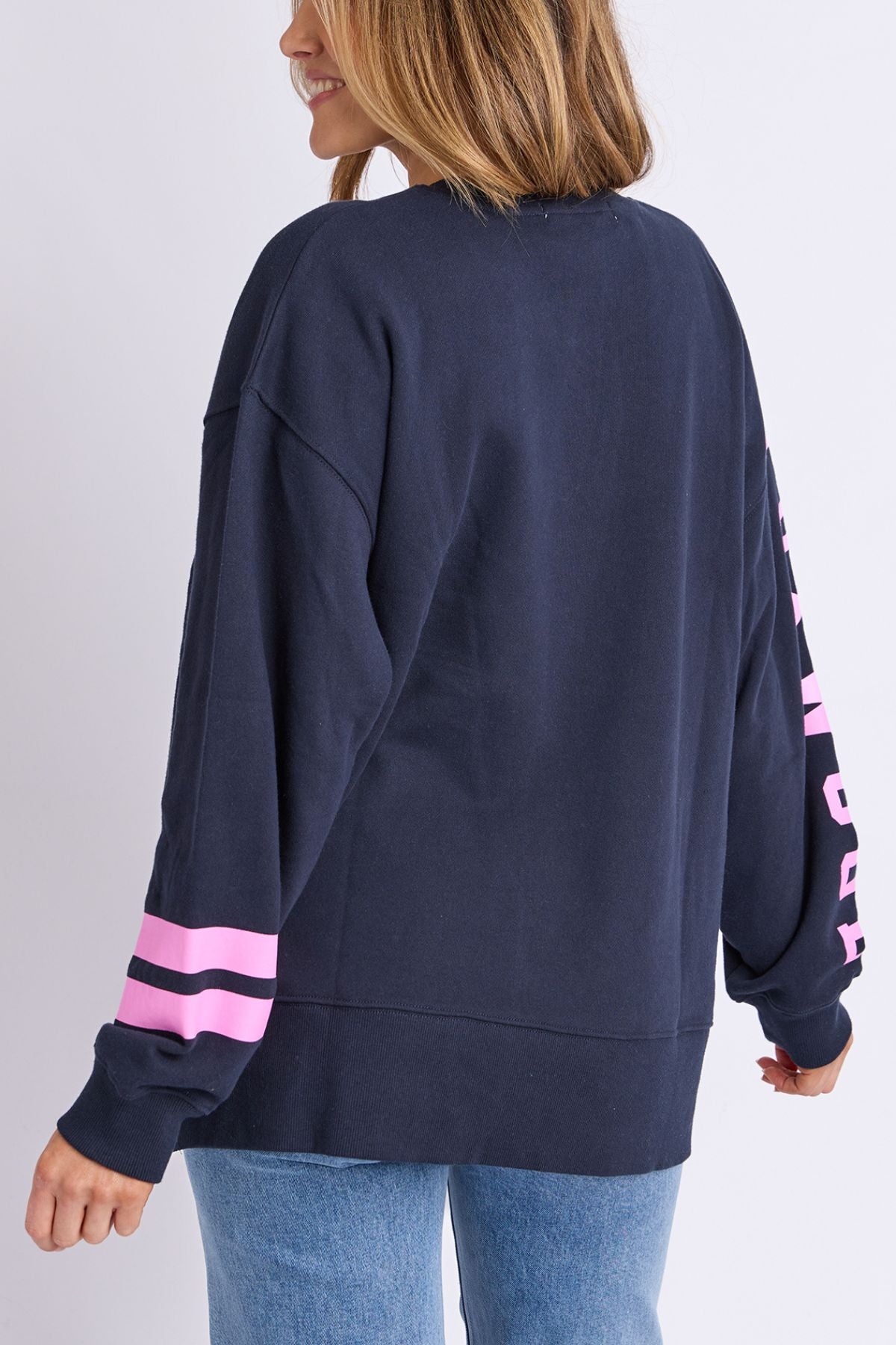 A woman with light brown hair wears the Foxwood Origin Crew, a relaxed-fit navy sweatshirt with pink stripes and sleeve text, styled with light blue jeans. She smiles, facing away from the camera for a perfect LeisureFit look.
