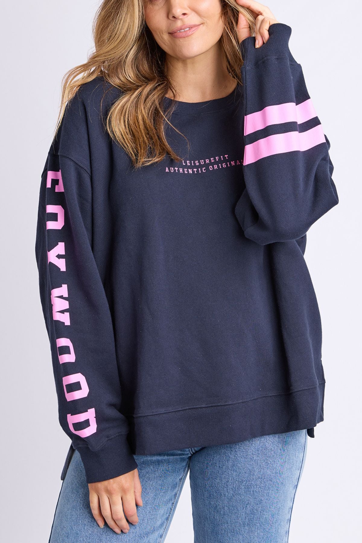 A woman with long, wavy hair wears a navy Foxwood Origin Crew featuring pink stripes and “HOLLYWOOD” in pink on the sleeve. She pairs this relaxed-fit fleece with light blue jeans and touches her hair.