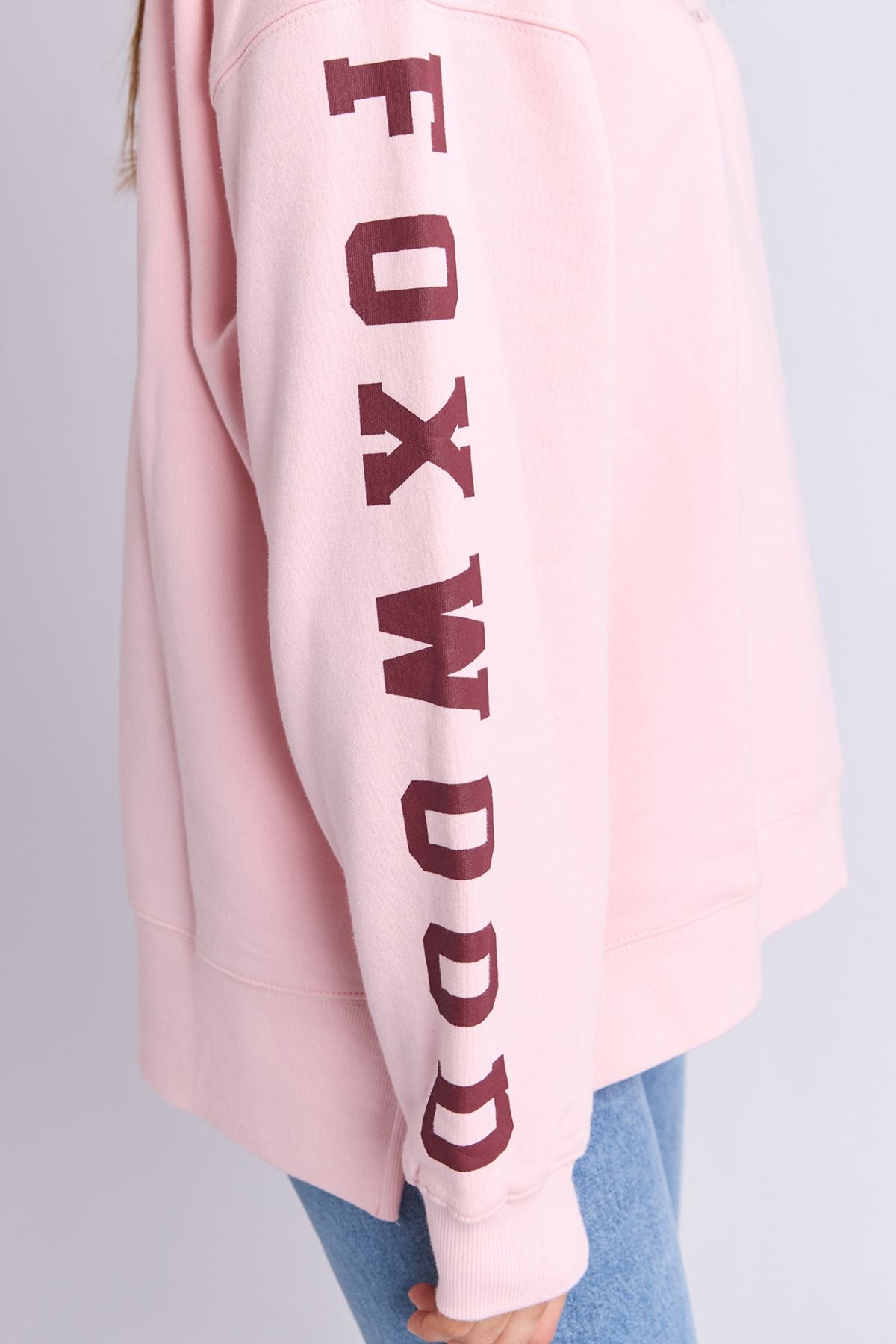 Someone wears the Foxwood Origin Crew, a relaxed-fit fleece in light pink with "FOXWOOD" boldly printed vertically in maroon on the sleeve, styled with light blue jeans.