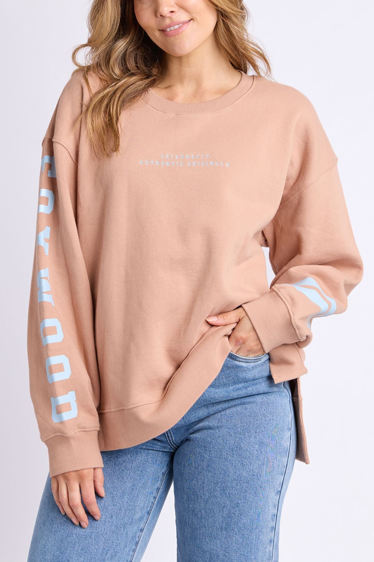 A woman wears the Foxwood Origin Crew in light brown, an oversized relaxed-fit fleece featuring white chest text and blue sleeve text, paired with light blue jeans, posing with one hand in her pocket for a modern look.