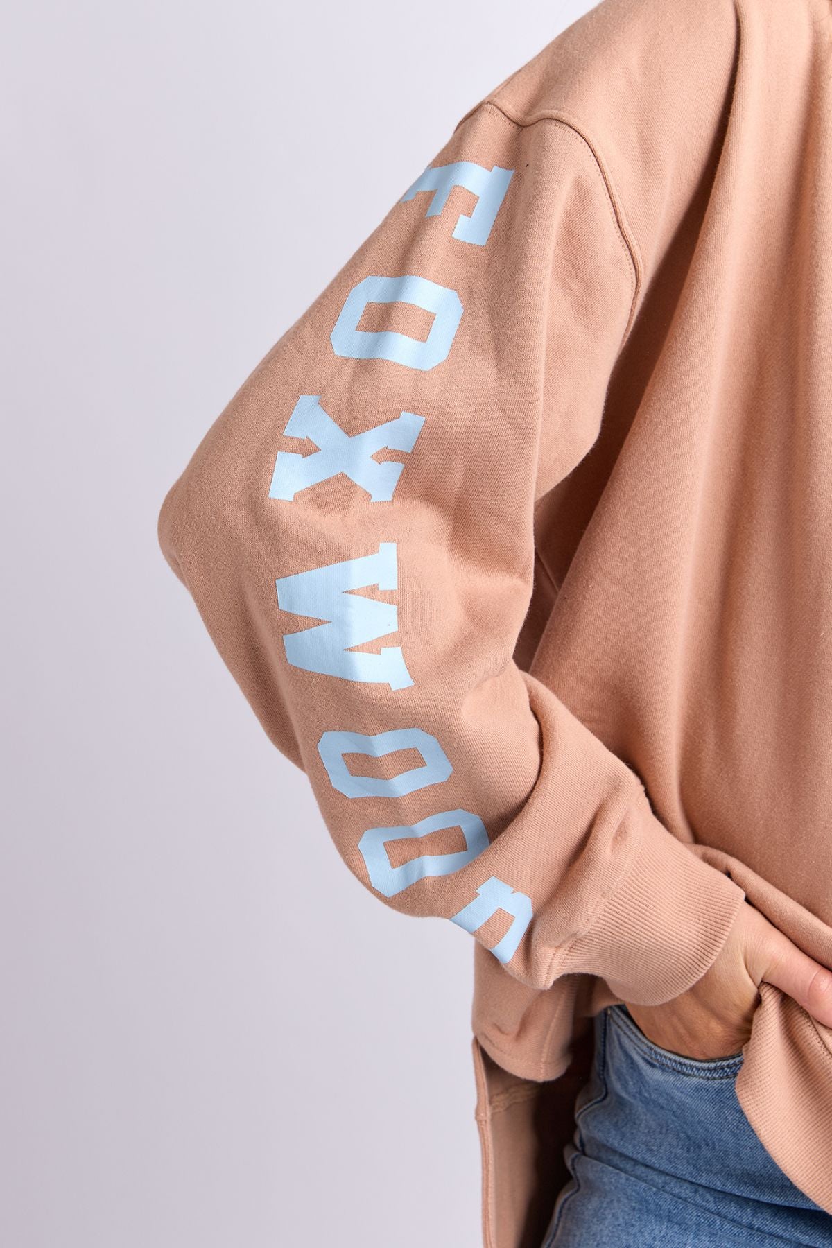 Wearing the Foxwood Origin Crew, a peach relaxed-fit fleece with bold light blue "FOXWOOD" lettering on the sleeve, styled with blue jeans for a modern look. Their arm bends as their hand rests in their pocket.
