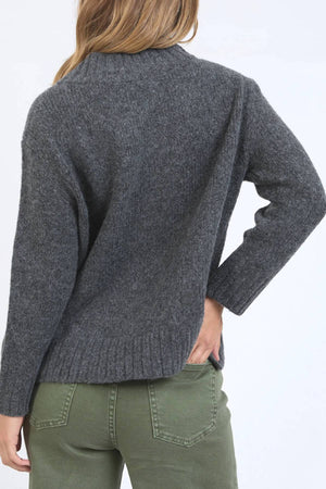 Someone with long hair is shown from the back against a plain light background, wearing the Foxwood Pepper Knit in dark gray and olive green pants, with one hand on their hip to showcase the sweater’s relaxed fit and cozy texture.