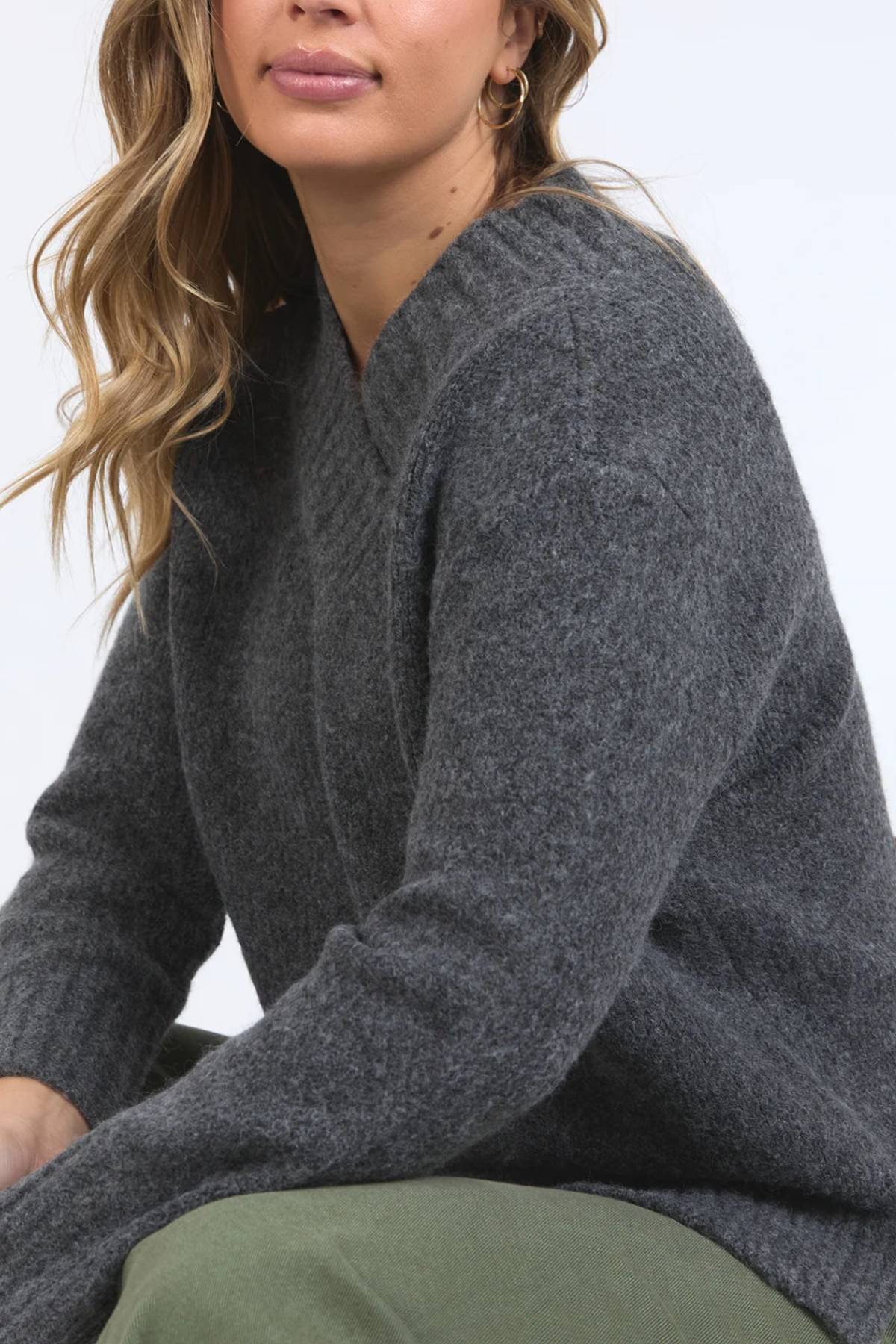A woman with wavy blonde hair sits against a light background, wearing the Foxwood Pepper Knit sweater in dark gray and olive green pants. Her face is partially visible, and she accessorizes with gold hoop earrings.
