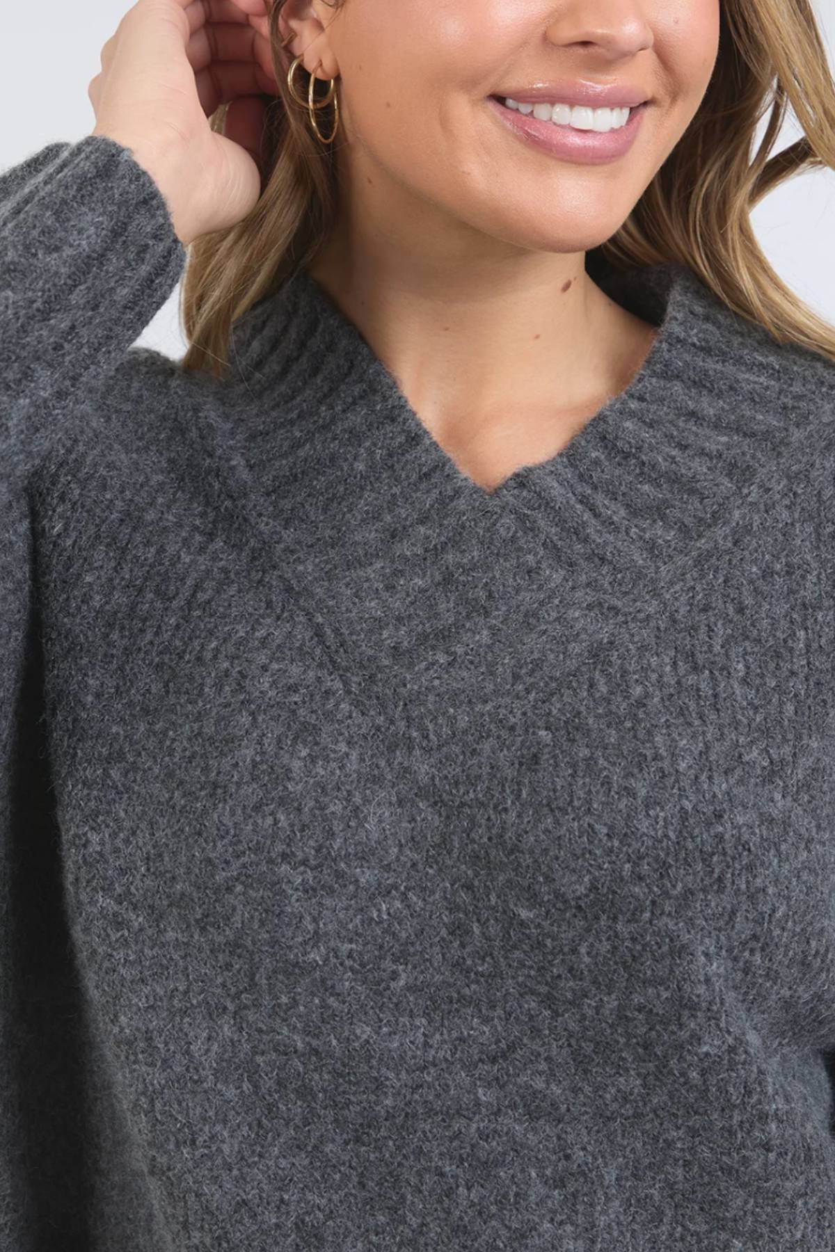 A woman smiles in the Foxwood Pepper Knit, a textured dark gray sweater featuring a relaxed fit and V-neckline, with gold hoop earrings and light pink lipstick, her hand lifted to her hair against a plain white background.