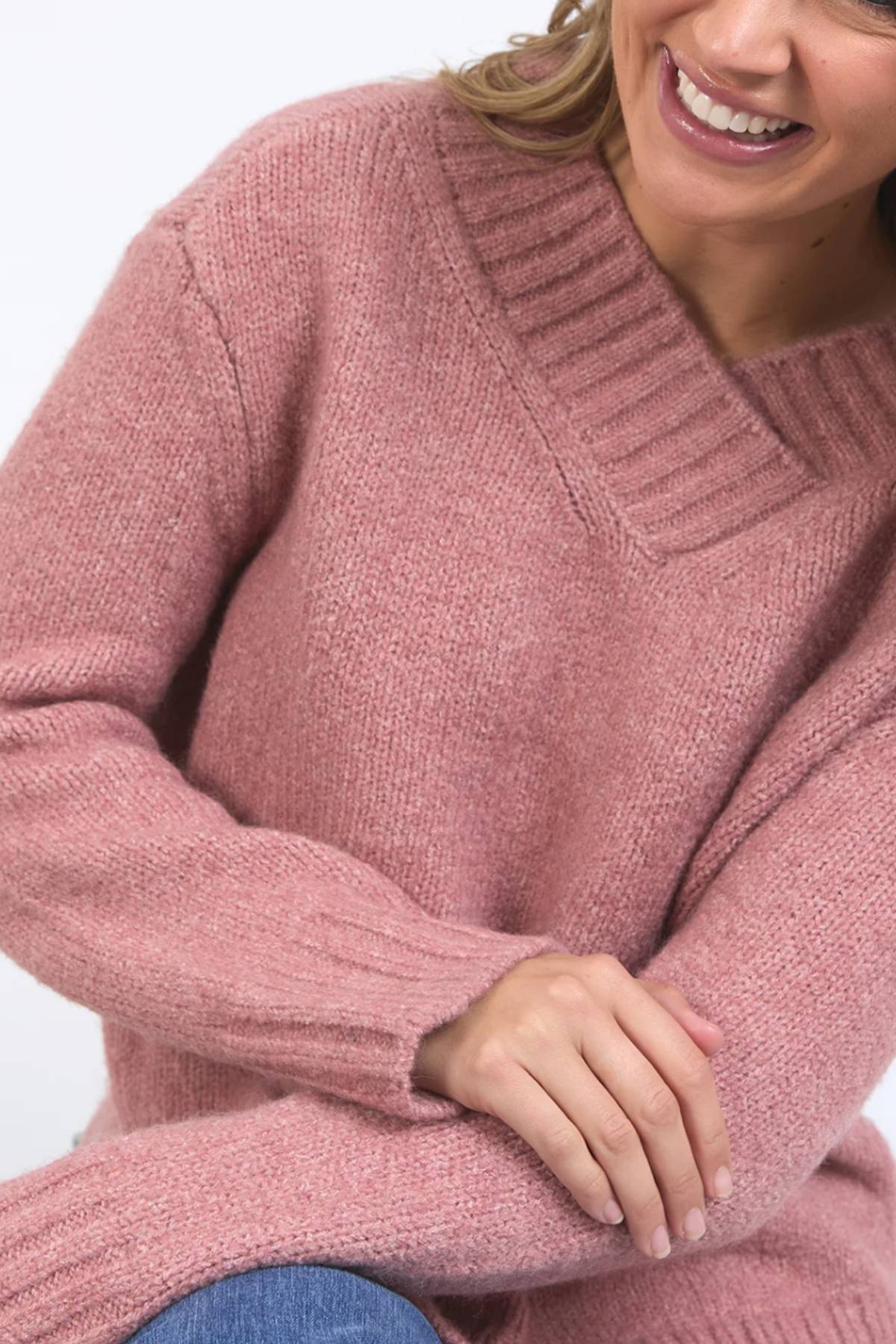 A person wearing the Foxwood Pepper Knit sweater in pink, featuring a cozy blend with ribbed neckline and sleeves, smiles with arms crossed against a plain background. Only their lower face and upper body are visible.