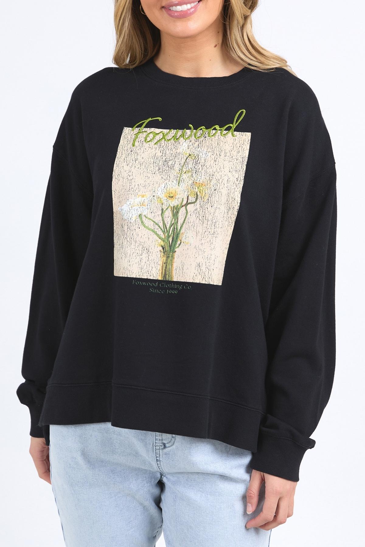 A woman wears the Foxwood Posy Crew, a black sweatshirt with yellow and white floral graphics and "Forward" in green script, paired with light blue jeans. For sizing, check the half chest measurement or contact customer service.