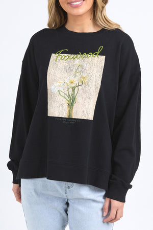 A woman wears the Foxwood Posy Crew, a black sweatshirt with yellow and white floral graphics and "Forward" in green script, paired with light blue jeans. For sizing, check the half chest measurement or contact customer service.