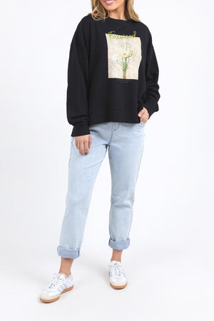 A person models the Foxwood Posy Crew, a black sweatshirt with a floral graphic, paired with light blue cuffed jeans and white sneakers against a plain white background. For sizing, see our half chest measurement or contact customer service. Head not fully visible.