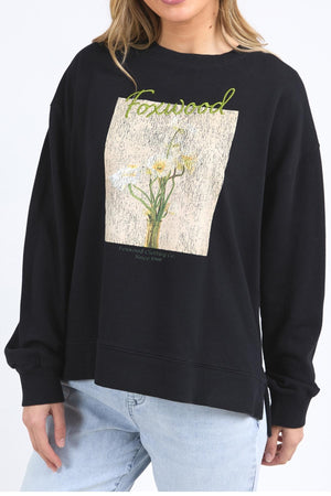 A person wears the Foxwood Posy Crew—a black relaxed-fit sweatshirt with a floral graphic and "Foxwood" in green—paired with light blue jeans. For the right size, check the half chest measurement or contact customer service.