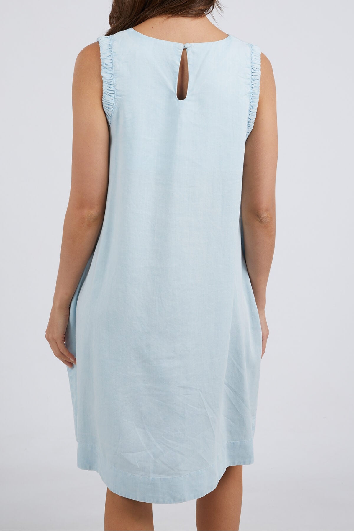 A woman is seen from the back wearing the sleeveless Foxwood Quinn Dress in light blue, featuring a relaxed fit, keyhole upper back opening, and ruffled armholes.