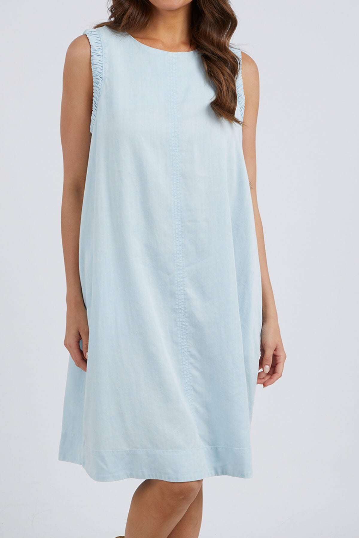 A woman models the Foxwood Quinn Dress, a sleeveless, relaxed-fit design with light blue fabric and subtle ruffle details on the shoulder seams, standing against a plain white background.