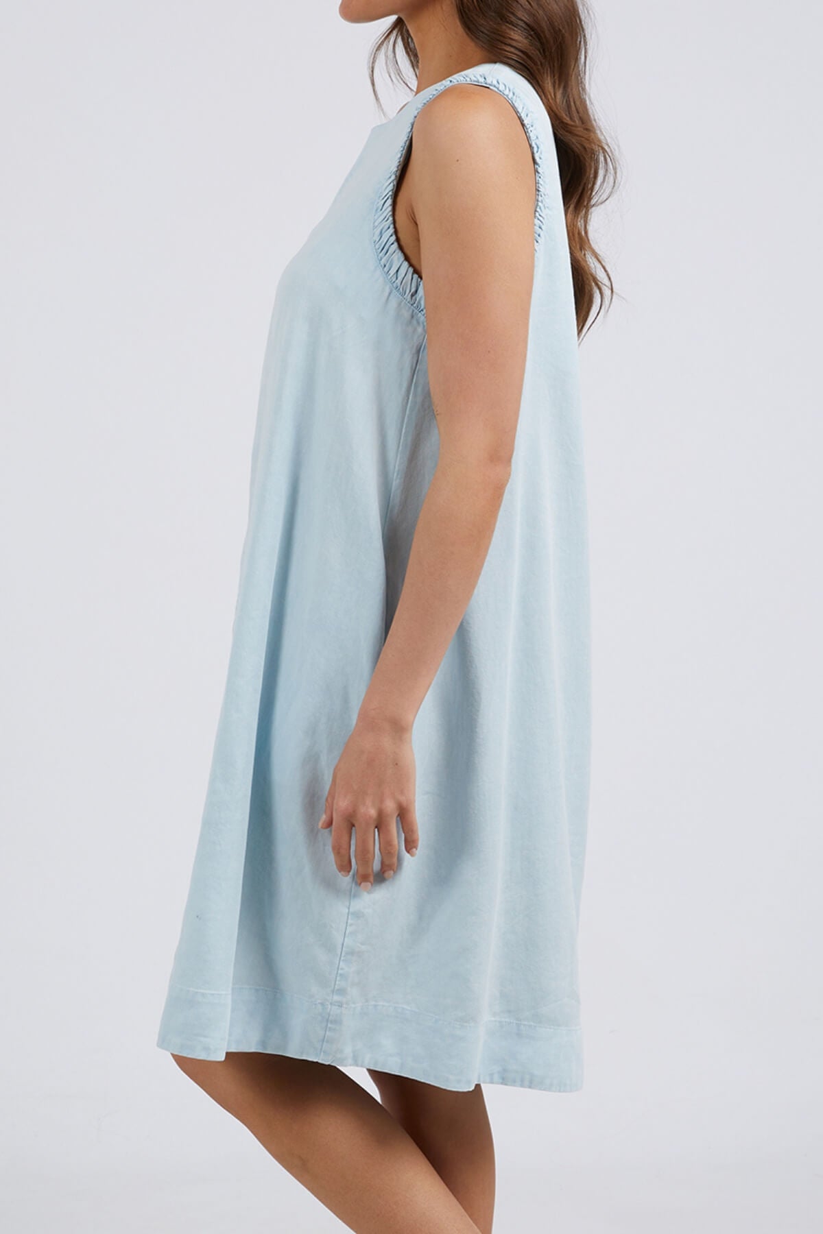 A woman models the Foxwood Quinn Dress—a light blue, sleeveless style with a relaxed fit and gathered shoulder details, made from a soft linen lyocell blend—standing sideways against a white background.