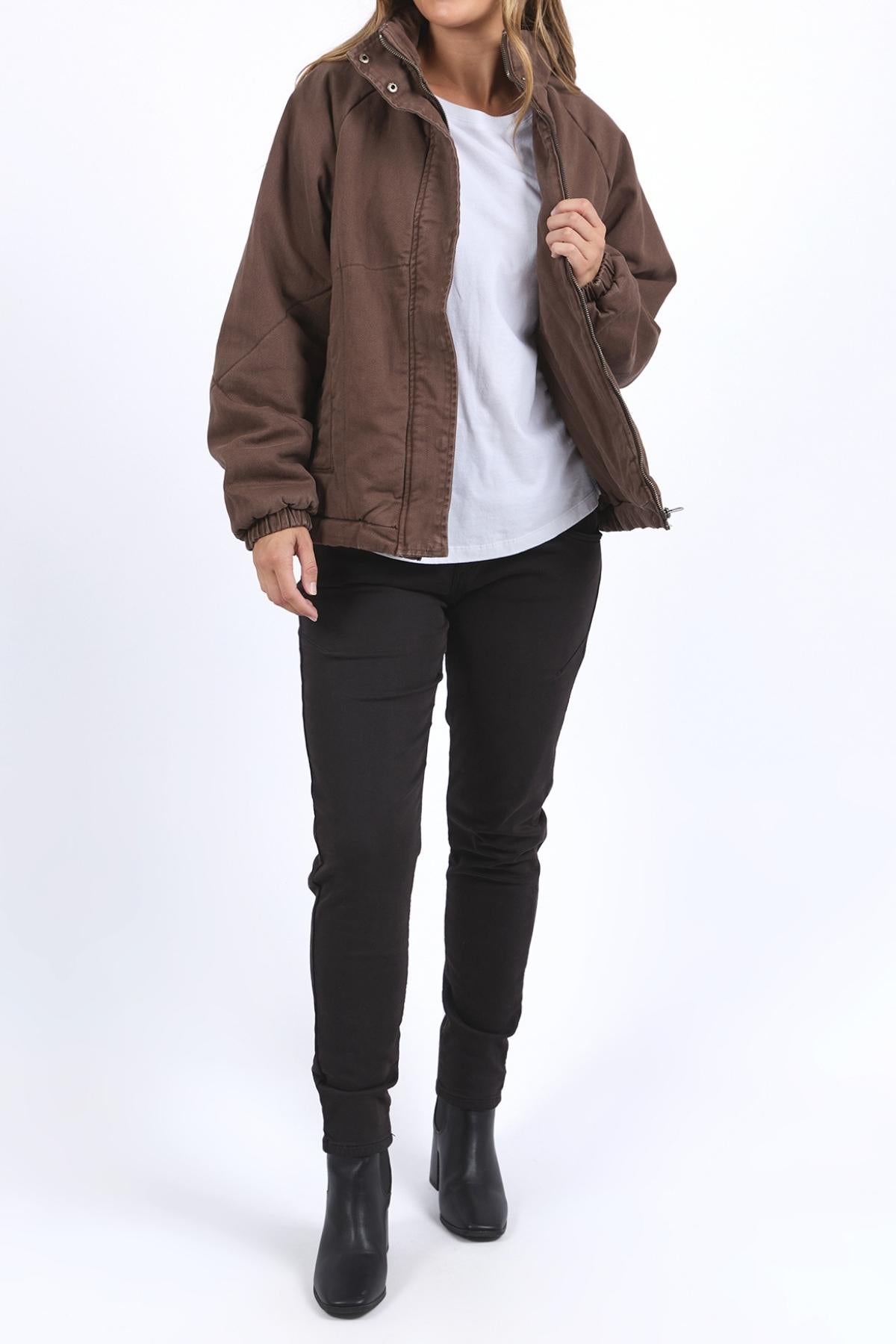 A person with loose hair stands against a white background wearing a Foxwood Rosalee Jacket in a cropped slouch style, partially unzipped over a white t-shirt, paired with black pants and black ankle boots.