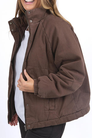 A person wears the Foxwood Rosalee Jacket in brown, cropped with a slouchy design, partially unzipped over a white shirt and dark pants, smiling as they turn their head slightly to the side.