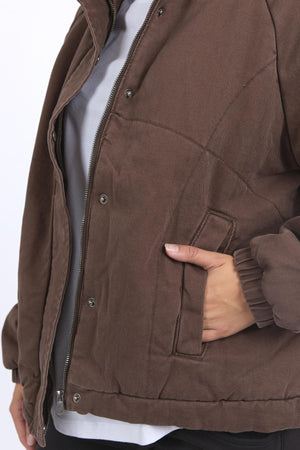 A person wears the Foxwood Rosalee Jacket, a brown, padded, cropped slouch design with a zipper and snap buttons. Their right hand is in the pocket, and the jacket is slightly open to reveal a white shirt underneath.