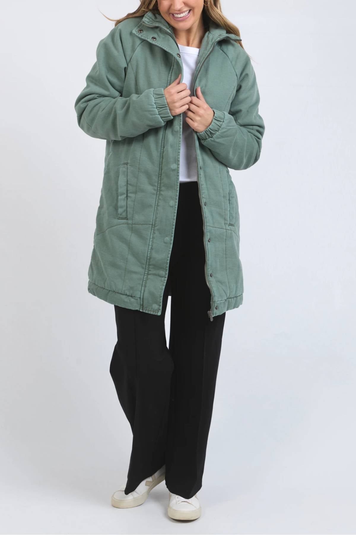 A woman wearing the Foxwood Rosalee Longline Jacket in light green over a white shirt, black pants, and white shoes stands against a plain light gray background, smiling as she holds the jacket’s collar.