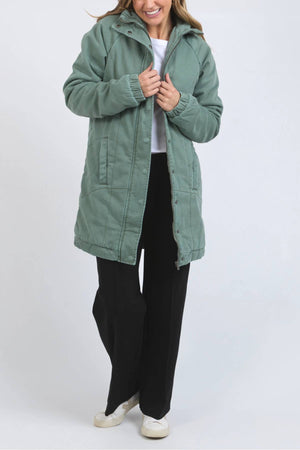 A woman wearing the Foxwood Rosalee Longline Jacket in light green over a white shirt, black pants, and white shoes stands against a plain light gray background, smiling as she holds the jacket’s collar.