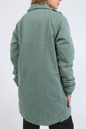 A person stands with their back to the camera against a plain light background, wearing the Foxwood Rosalee Longline Jacket in sage green with visible seam lines and padded detailing.