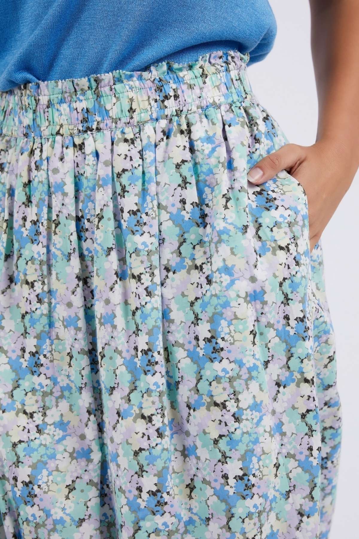 A person wearing a blue top and the high-waisted Foxwood Rosette Floral Skirt, featuring a vibrant light blue and green floral print, with their hand in the skirt’s pocket.