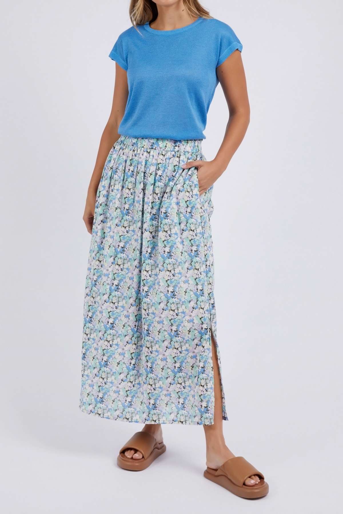 A woman wears a blue short-sleeve top and the Foxwood Rosette Floral Skirt, featuring a side slit and pockets. She pairs the look with tan sandals and stands against a plain background, one hand in her pocket.