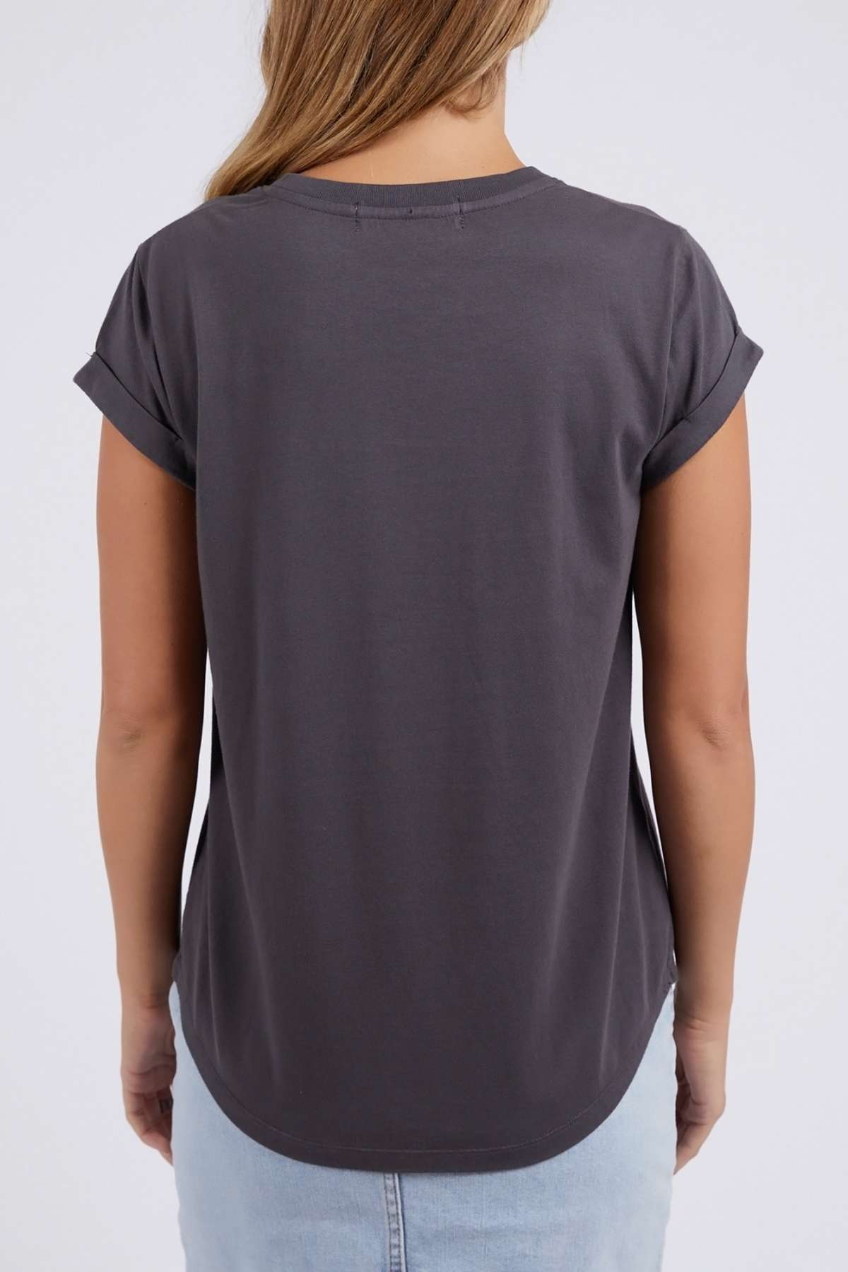 A woman with light brown hair wears the Foxwood Rosette Tee in dark gray, featuring short sleeves and a relaxed fit, paired with a light blue skirt. She is shown from the back against a plain white background.