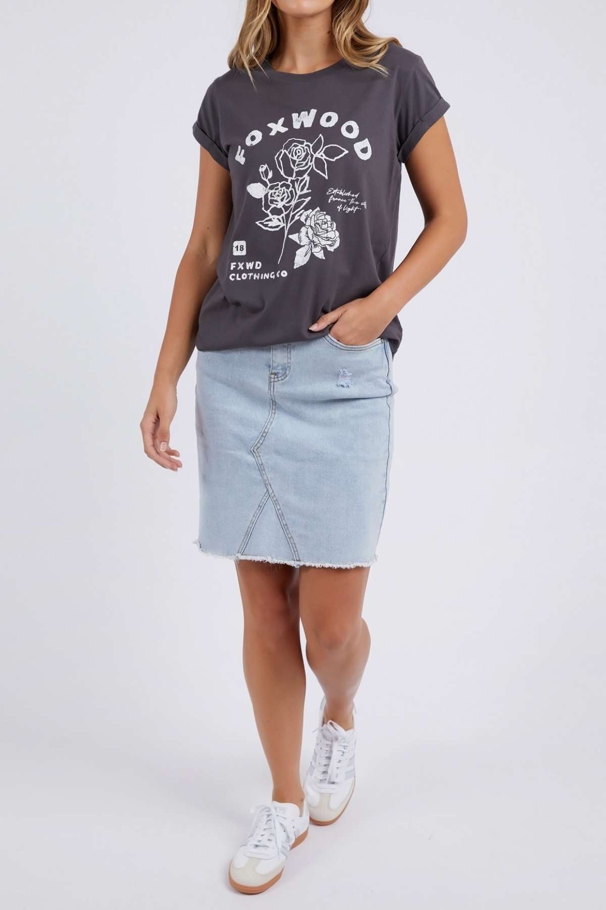 A woman models the Foxwood Rosette Tee, a dark graphic t-shirt featuring white floral designs and lettering, styled with a light blue denim skirt and white sneakers against a plain backdrop.