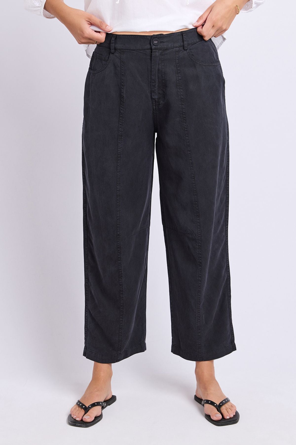 A person wearing the Foxwood Roy Pant in black and black sandals stands against a plain light background. The image captures the pants from the waist down, with their hands resting on the waistband.
