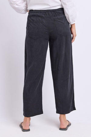 A person wearing the Foxwood Roy Pant in black, paired with a white long-sleeve blouse and black flip-flops, photographed from the back against a plain background.