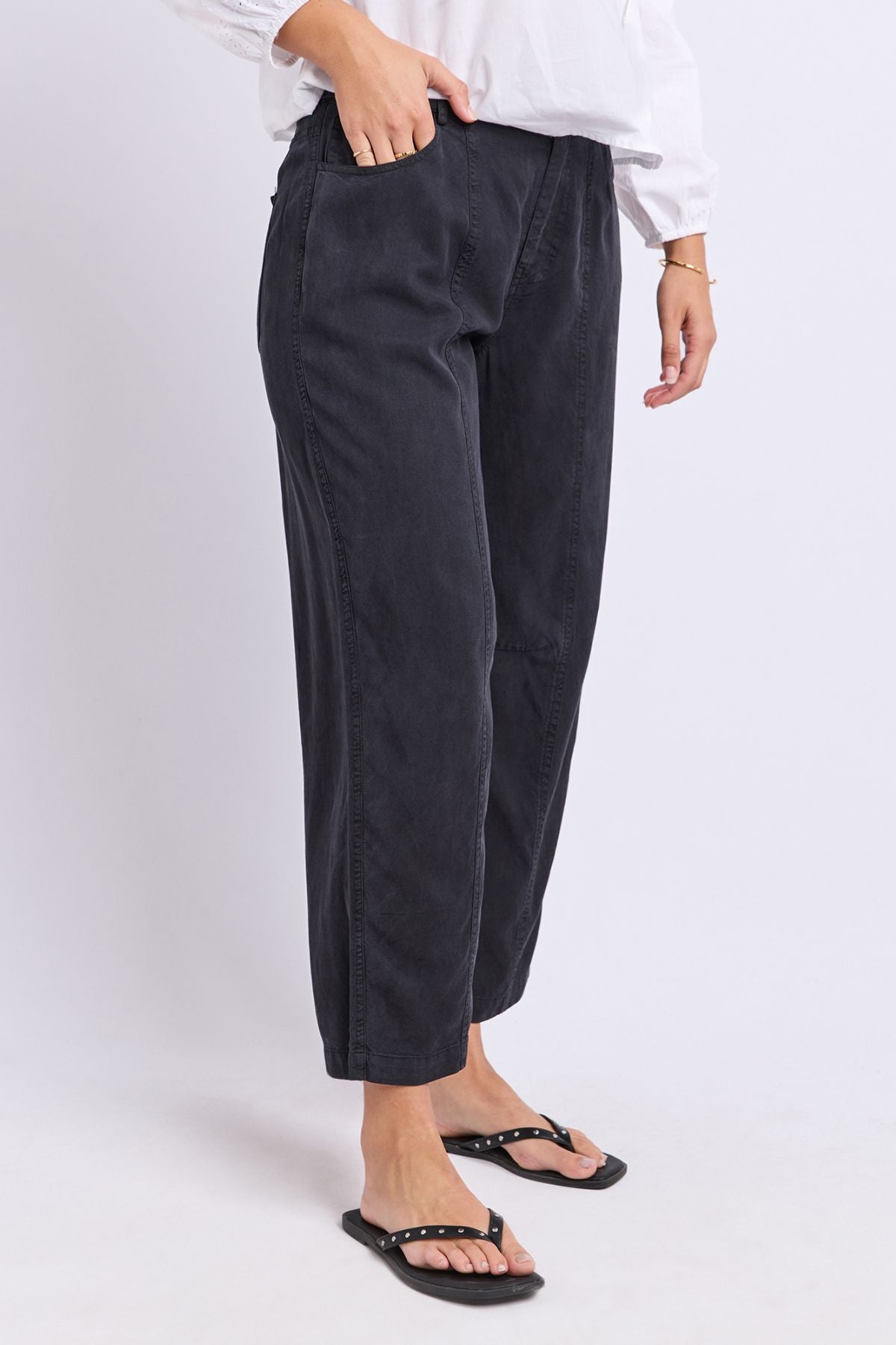 A person wearing the Foxwood Roy Pant, paired with a white long-sleeve blouse and black studded sandals, stands against a plain white background with one hand in their pocket.